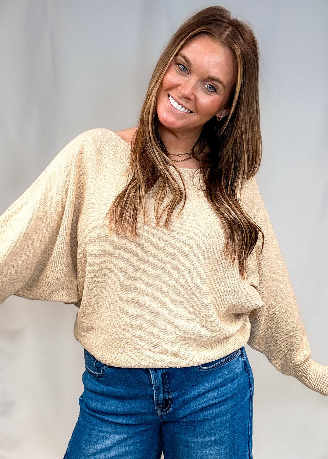A smiling woman wearing a beige knit sweater with wide batwing sleeves and blue jeans, standing against a light gray studio backdrop.