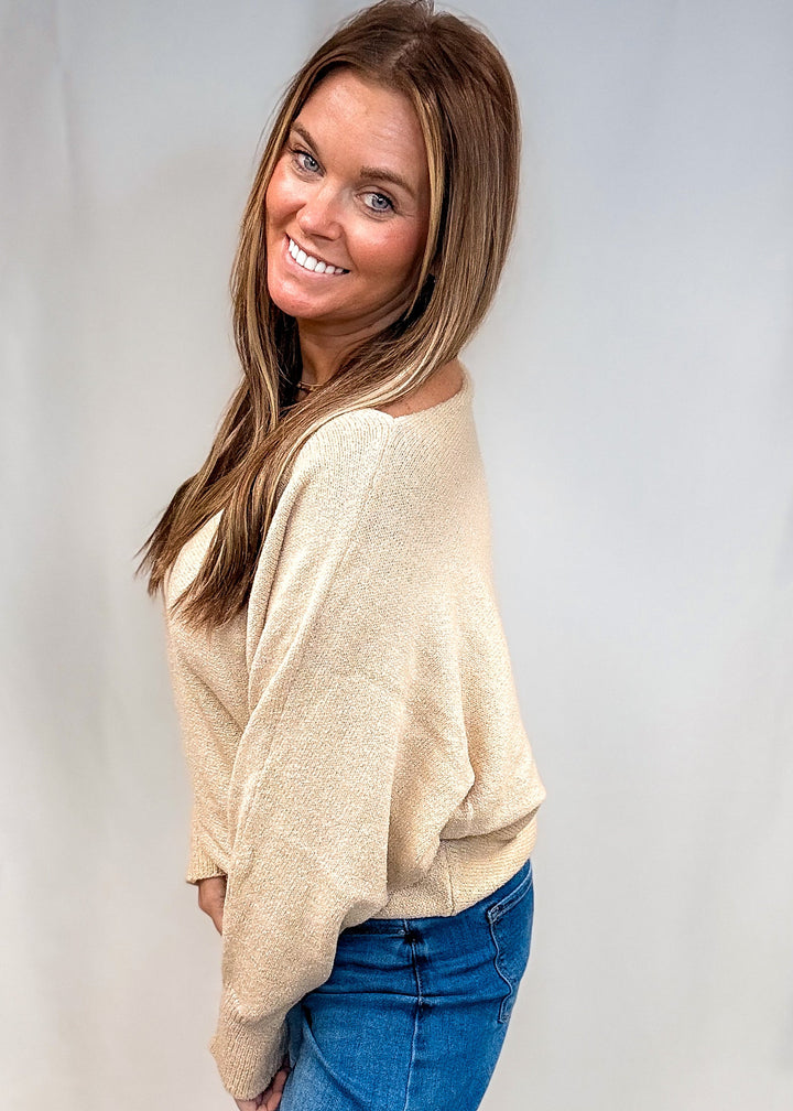 A woman modeling a beige knit sweater with drop shoulders and blue jeans, standing against a white studio backdrop.