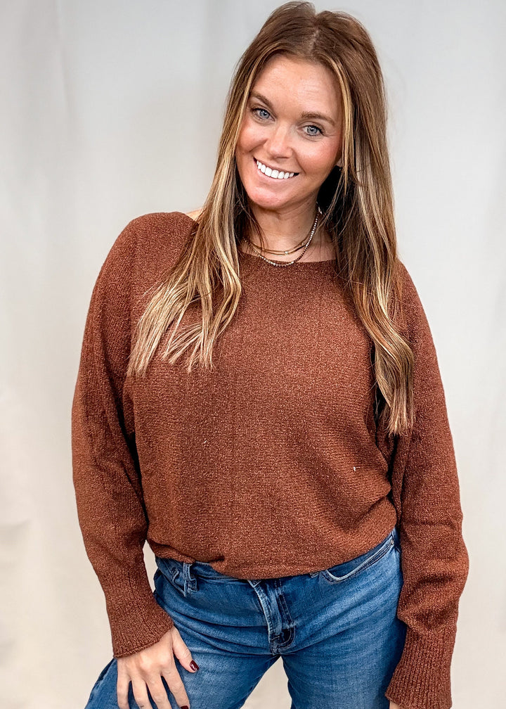 A smiling woman wearing a textured brown knit sweater and blue jeans, standing against a light neutral studio backdrop.