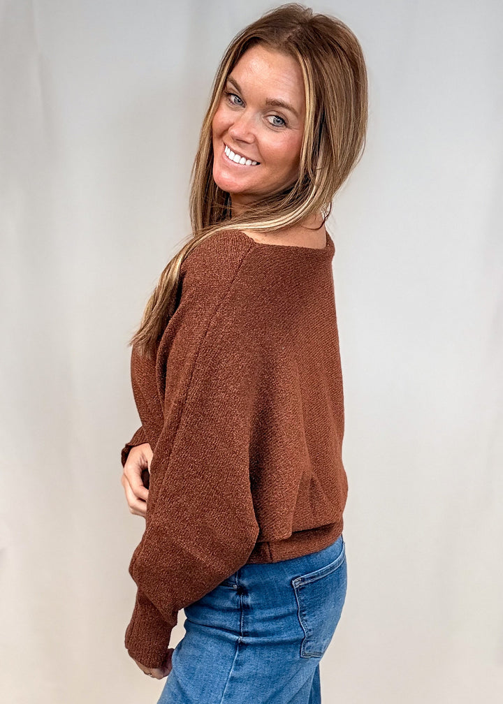 A woman wearing a brown fuzzy knit sweater and blue jeans, photographed from the back-three-quarter angle against a white studio backdrop.