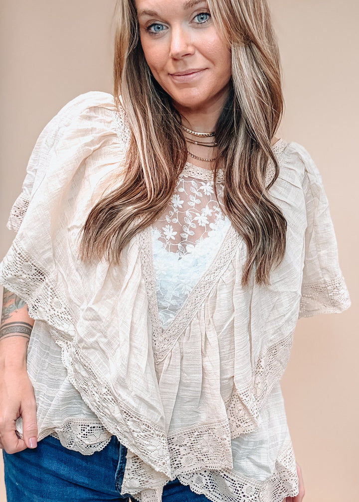 A woman wearing a cream-colored, semi-sheer ruffled lace top over a light blue floral lace top, paired with blue jeans, against a solid beige background.