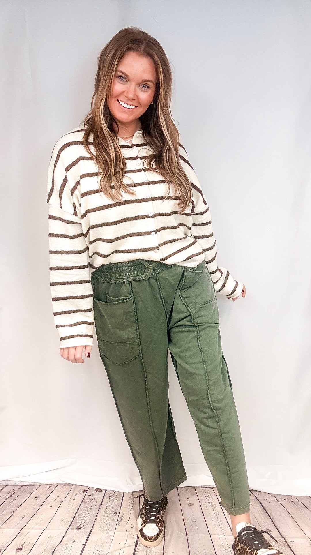 Woman wearing green barrel sweatpants with white and brown cardigan on white background