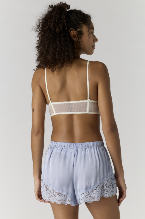 Woman wearing a white lace bralette and light blue shorts on a plain background