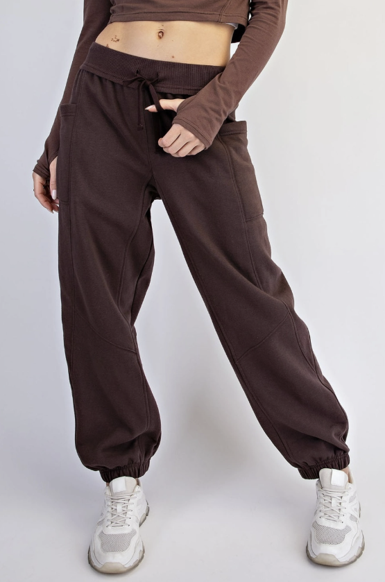 A person wearing dark brown jogger sweatpants with a drawstring waist, paired with a matching brown long-sleeve crop top and white sneakers, shown against a light gray studio background.