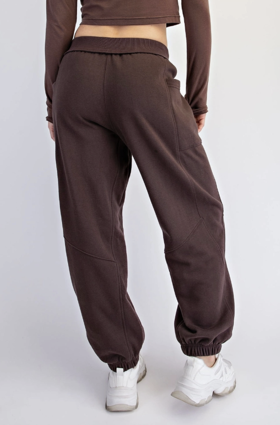 A person shown from the back wearing loose brown jogger sweatpants with elastic cuffs, paired with white sneakers, against a light gray studio background.