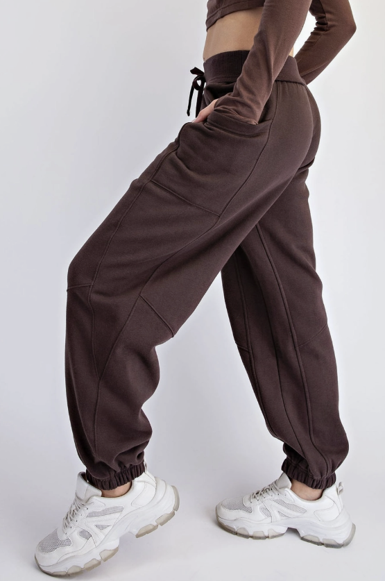 A person wearing dark brown jogger sweatpants with elastic cuffs, paired with white sneakers, shown from the waist down against a plain light gray studio background.