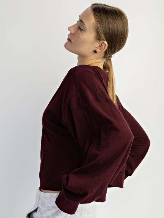 A woman wearing a dark burgundy long-sleeve cropped sweatshirt and light gray sweatpants, shown from a side profile against a plain light gray studio background.
