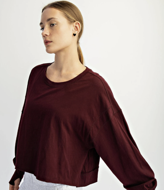 A woman wearing a dark burgundy long-sleeve crop top and light gray sweatpants, photographed against a clean white studio background.