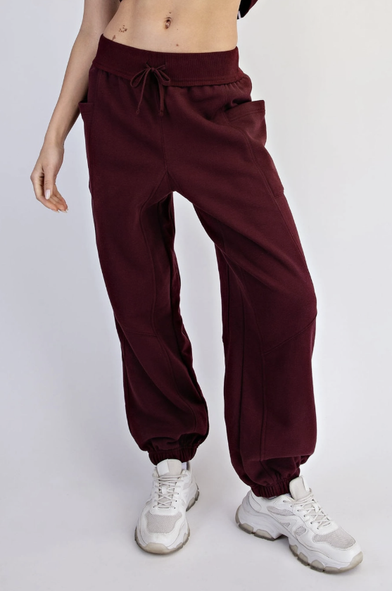 A person wearing loose burgundy jogger sweatpants with elastic cuffs and white chunky sneakers, photographed against a plain light gray studio background.