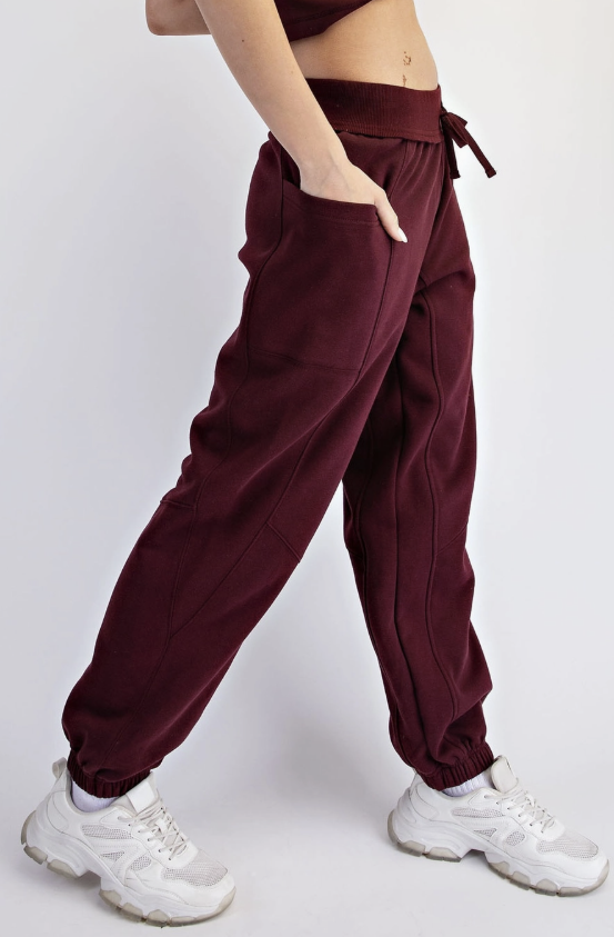 A person wearing burgundy jogger sweatpants with a drawstring waist and elastic cuffs, paired with white sneakers, shown from the waist down against a light gray studio background.