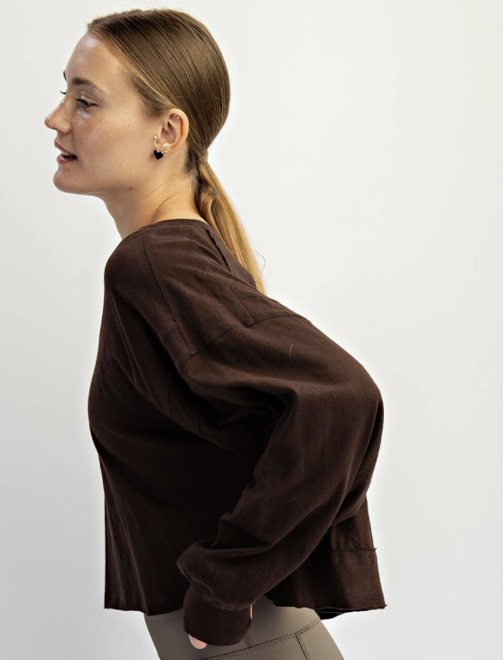 A woman in side profile wearing a dark brown cropped long-sleeve top and taupe leggings against a plain white studio background.