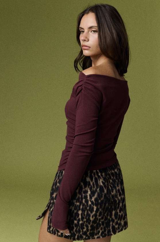 A woman wearing a dark burgundy off-the-shoulder long-sleeve top and a leopard-print mini skirt, photographed against a solid olive green studio background.