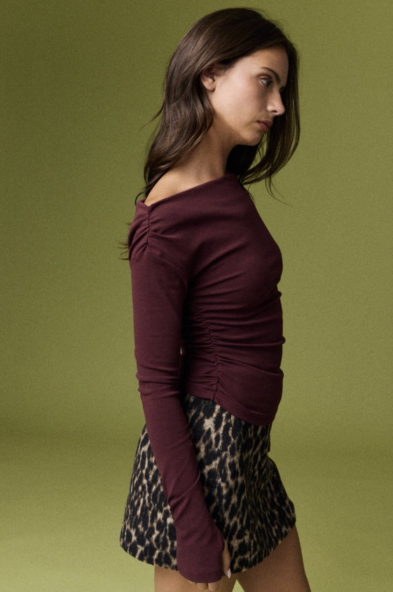 A woman in side profile wearing a ruched off-the-shoulder long-sleeve top in deep burgundy and a leopard-print skirt, set against a solid olive green studio background.