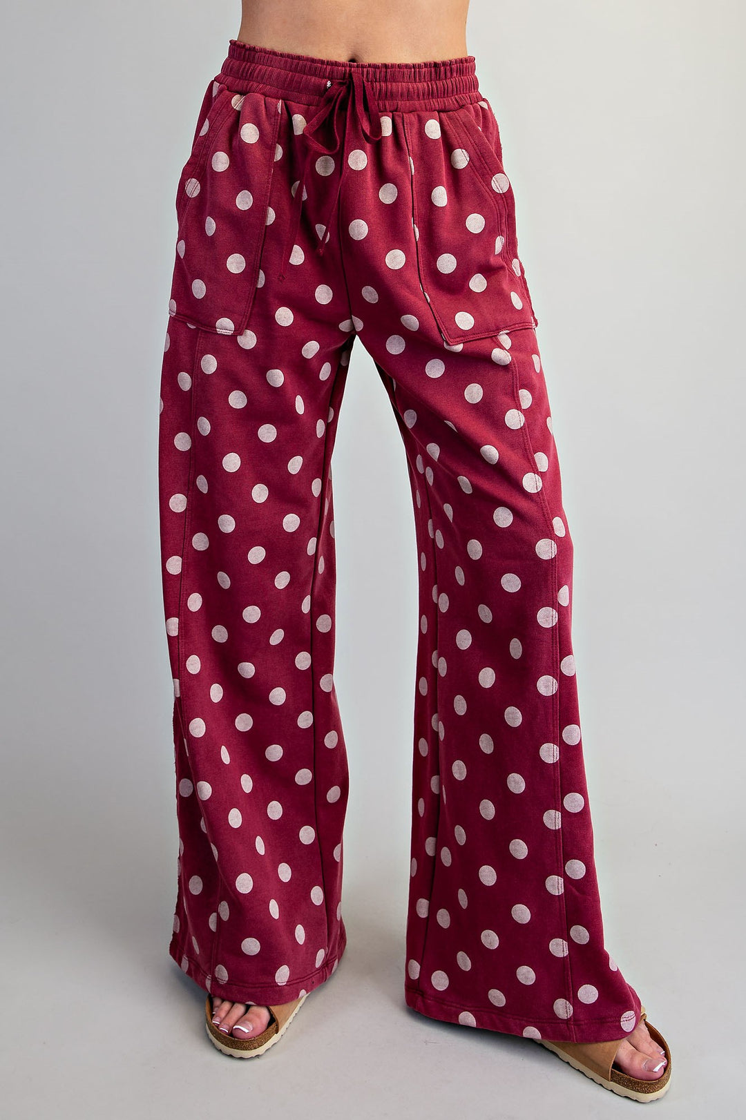 A person wearing wide-leg drawstring pants in burgundy with a white polka dot pattern, paired with pink sandals, shown from the waist down against a light gray studio background.