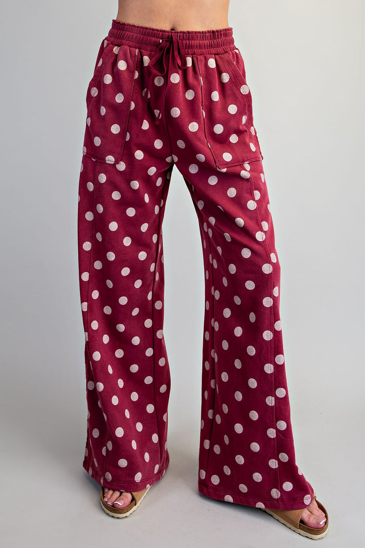 A person wearing wide-leg drawstring pants in burgundy with a white polka dot pattern, paired with pink sandals, shown from the waist down against a light gray studio background.