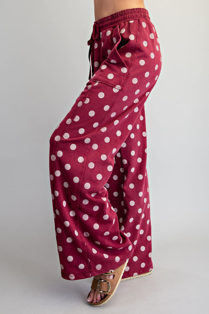 A person wearing wide-leg drawstring pants in burgundy with a white polka dot pattern, paired with brown leather slide sandals, shown from the waist down against a light gray studio background.
