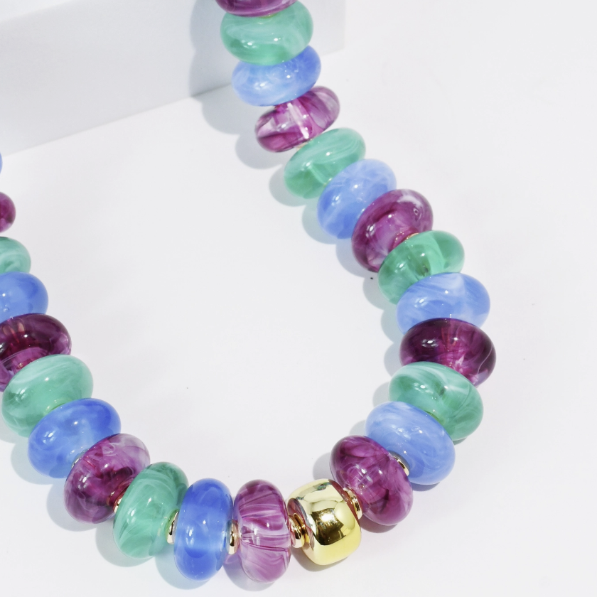 A beaded necklace made of glossy, translucent round beads in shades of purple, blue, and green, accented by a shiny gold cylindrical bead at the front, displayed on a clean white surface.