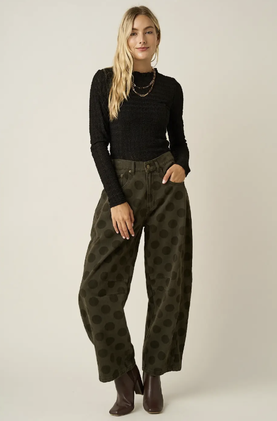 A woman modeling a black long-sleeve knit top and olive green wide-leg pants with a dark brown polka dot pattern, paired with dark brown ankle boots, against a plain light beige studio background.