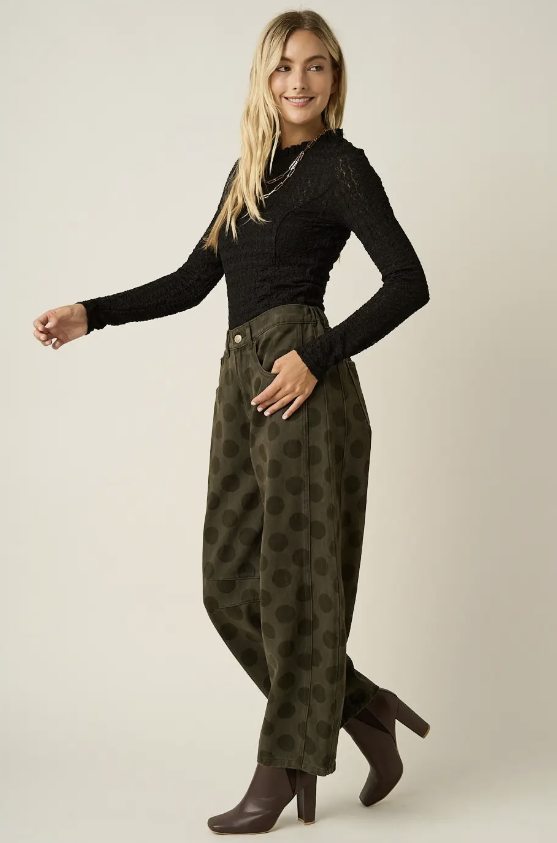A woman modeling a black long-sleeve lace-trim top and olive green wide-leg pants with a dark green polka dot pattern, paired with dark brown heeled boots, against a plain light beige studio background.