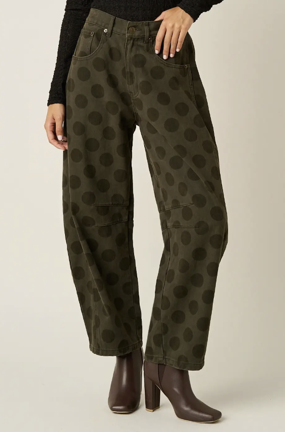 A person wearing olive green wide-leg pants with a dark brown polka dot pattern, styled with a black long-sleeve top and dark brown heeled ankle boots against a light beige studio background.