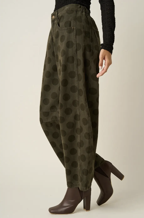 A person wearing olive green wide-leg pants with a dark brown polka dot pattern, styled with a black long-sleeve top and dark brown heeled ankle boots, shown from the waist down against a light beige studio background.