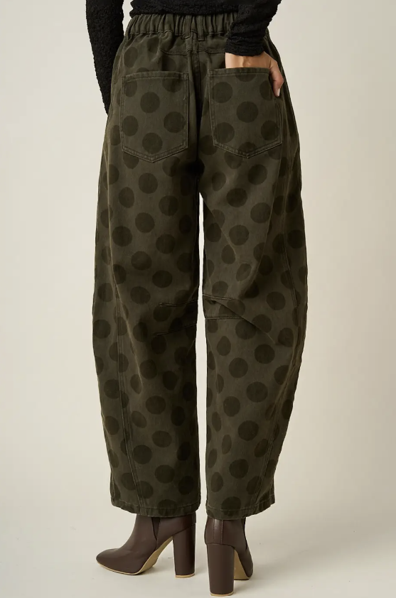 A person shown from the back wearing loose dark olive pants with a darker tonal polka dot pattern, a black long-sleeve top, and brown heeled ankle boots against a plain light beige studio background.