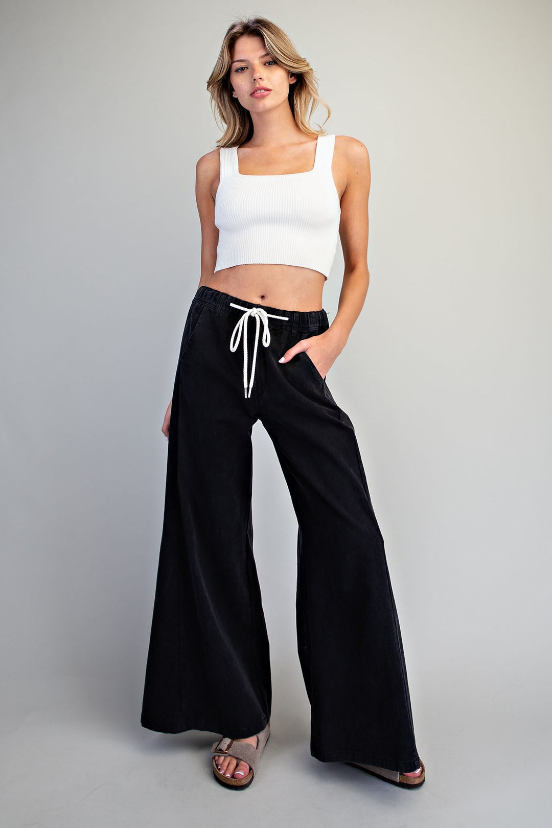 A woman modeling black wide-leg drawstring pants with a white crop top, standing against a light gray studio background.