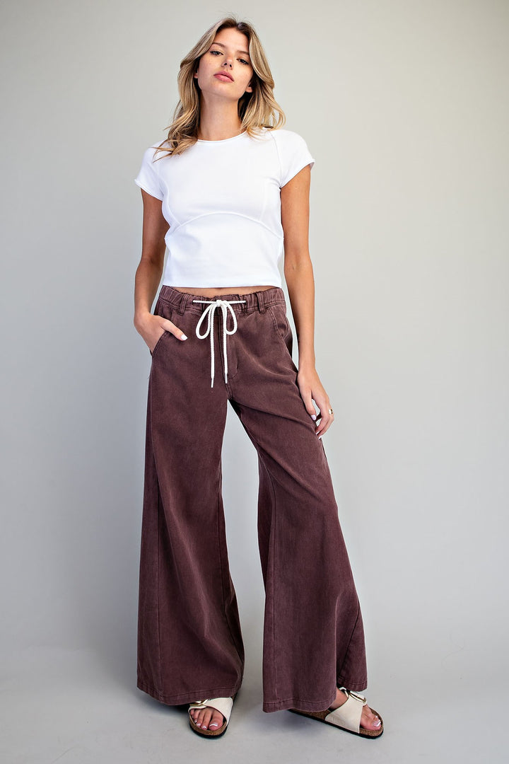A woman modeling a pair of wide-leg brown corduroy pants with a white drawstring, paired with a white t-shirt and white slide sandals, against a solid light gray background.