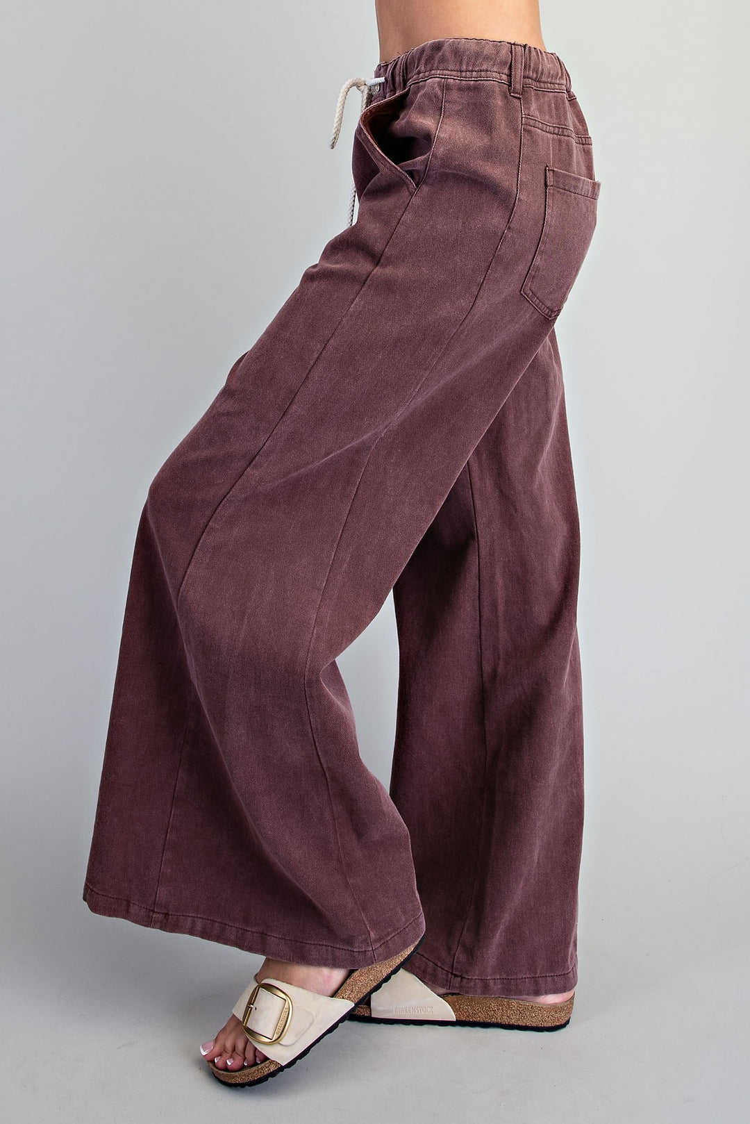 A person wearing wide-leg mauve corduroy pants with a drawstring waistband, paired with beige slide sandals, shown from the waist down against a light gray studio background.