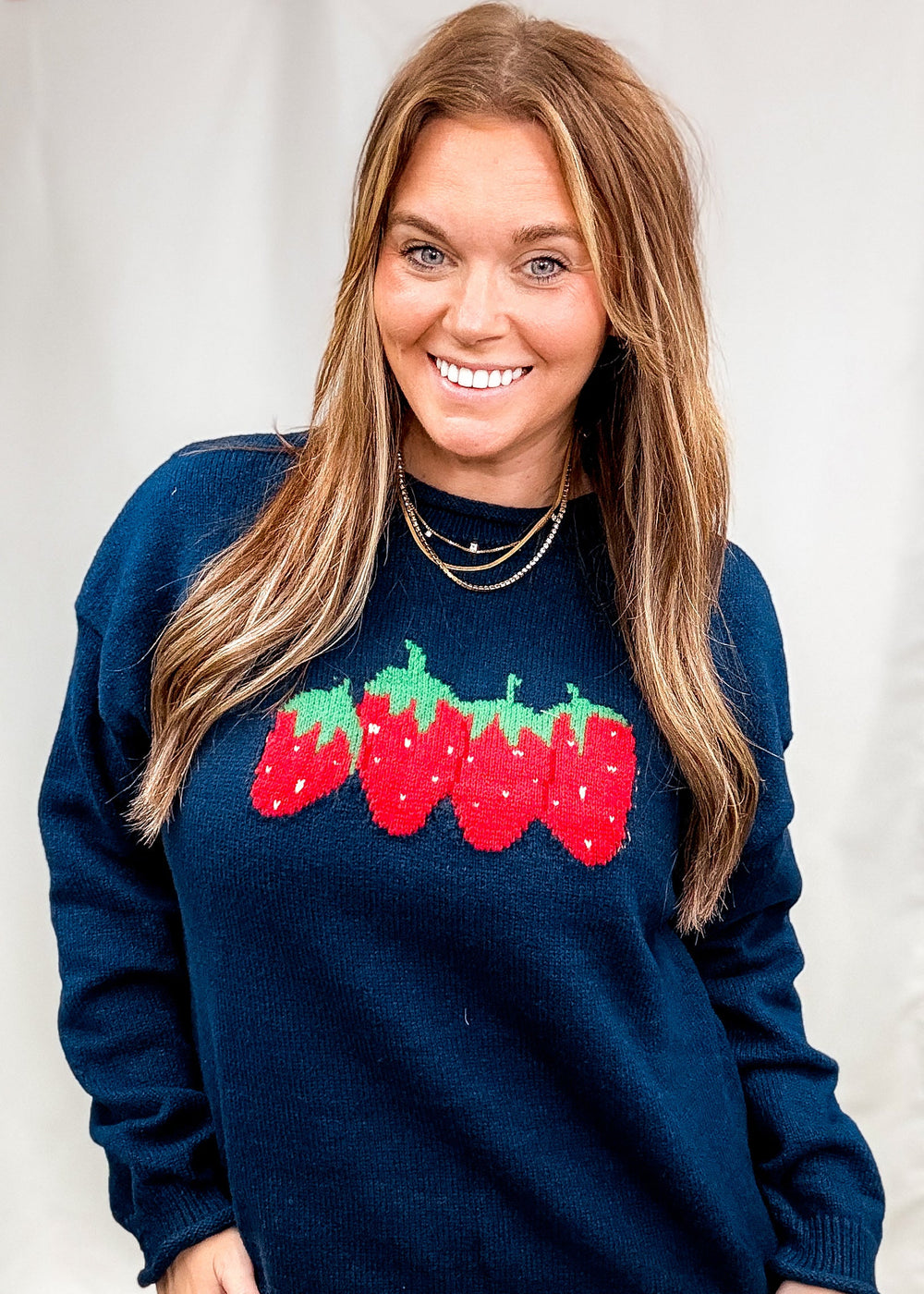 Woman wearing navy blue sweater with strawberry design on front with beige pants on plain background