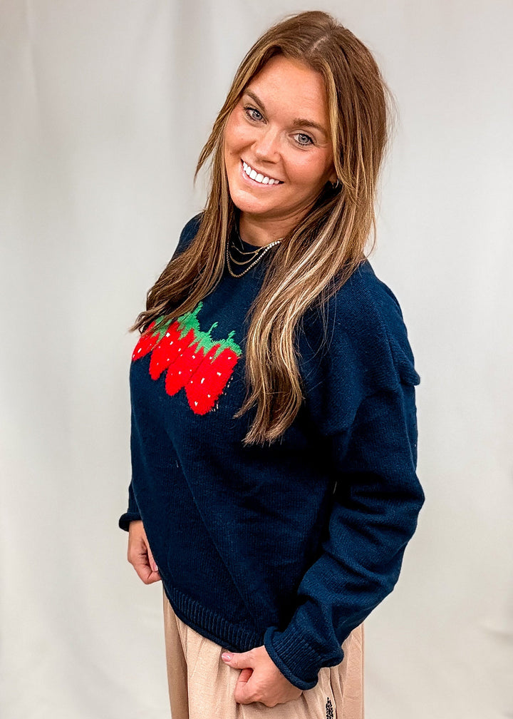 Woman wearing navy blue sweater with strawberry design on front with beige pants on plain background