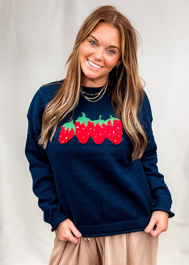 Woman wearing navy blue sweater with strawberry design on front with beige pants on plain background