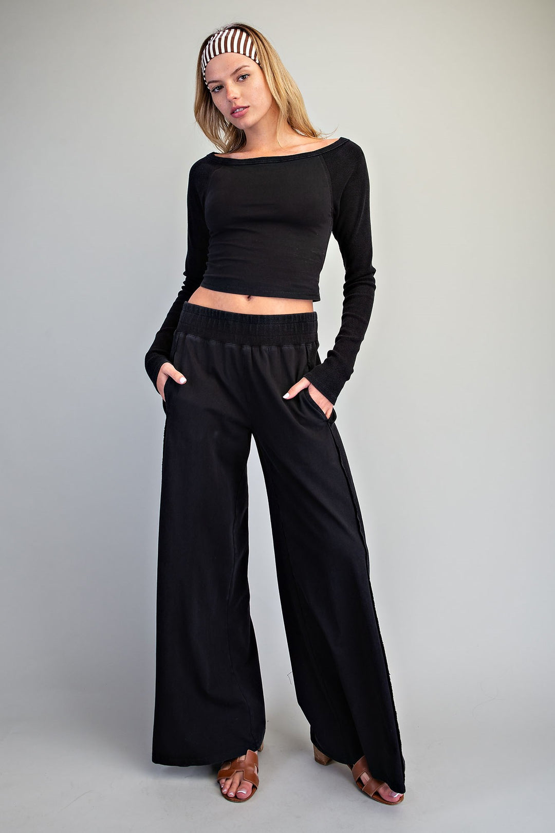 A woman modeling a black long-sleeve crop top and matching black wide-leg pants with side pockets, paired with brown heeled sandals and a black-and-white striped headband, against a light gray studio background.