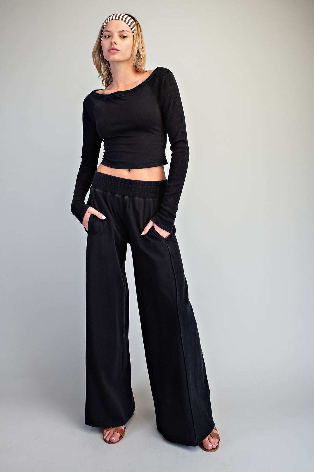A woman modeling a black long-sleeve cropped top and matching wide-leg pants against a plain light gray studio background.