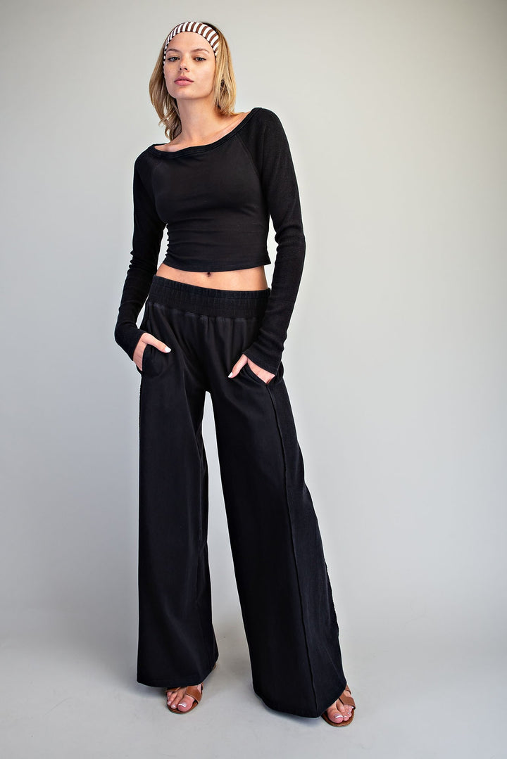 A woman modeling a black long-sleeve cropped top and matching wide-leg pants against a plain light gray studio background.