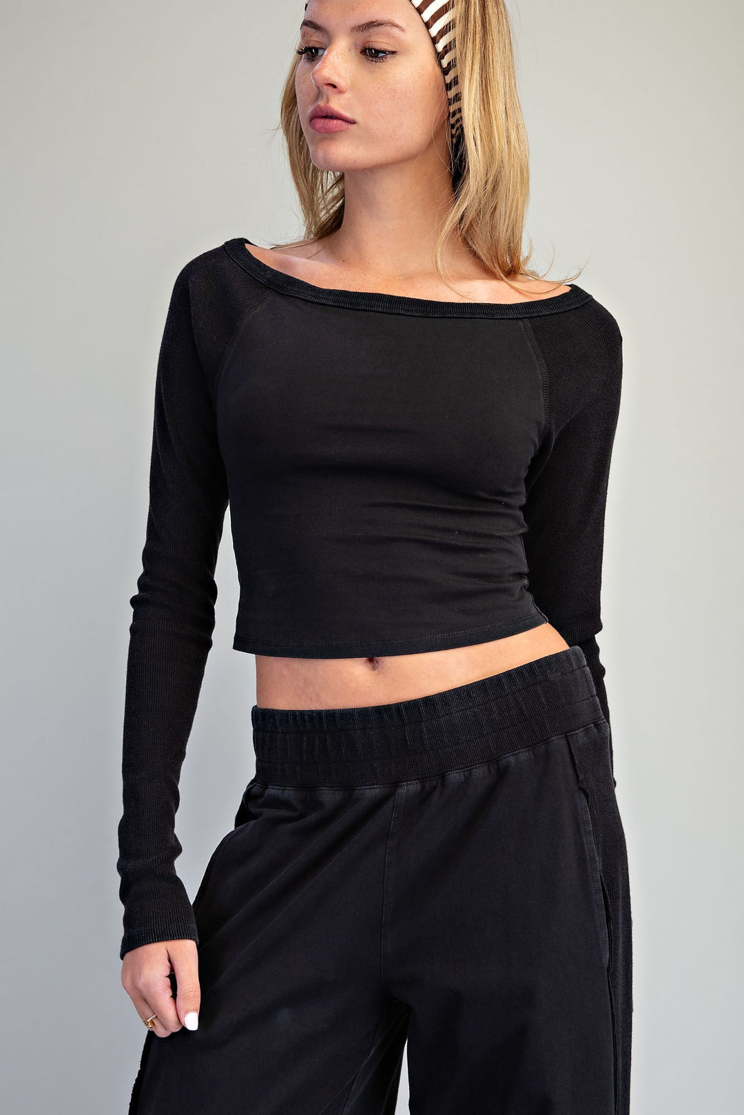 A woman wearing a black long-sleeve off-the-shoulder crop top and black wide-leg pants, photographed against a plain light gray studio background.