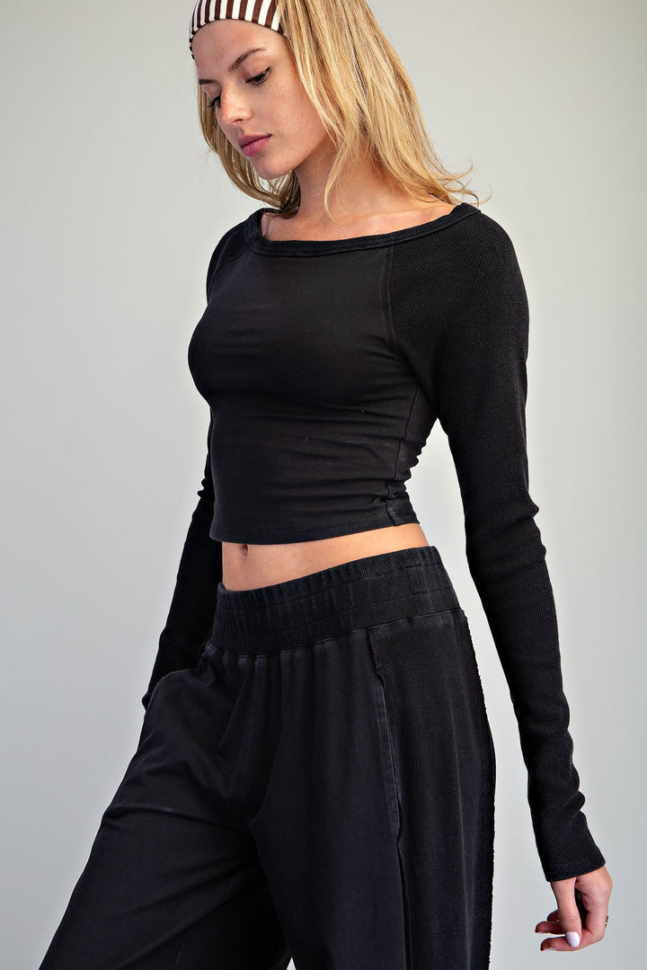 A woman wearing a black long-sleeve cropped top with a wide boat neckline and black wide-leg pants, photographed against a plain light gray studio background.