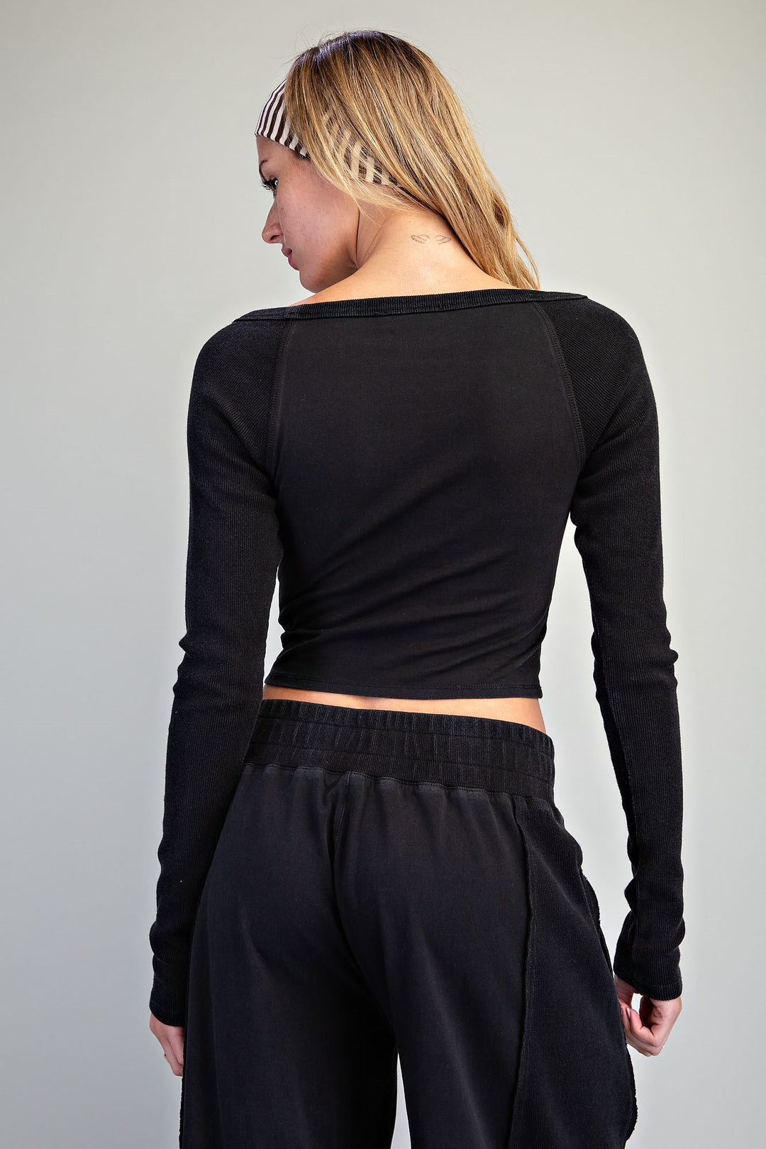 A woman shown from the back wearing a black long-sleeve crop top with raglan sleeves and black sweatpants against a plain light gray studio background.