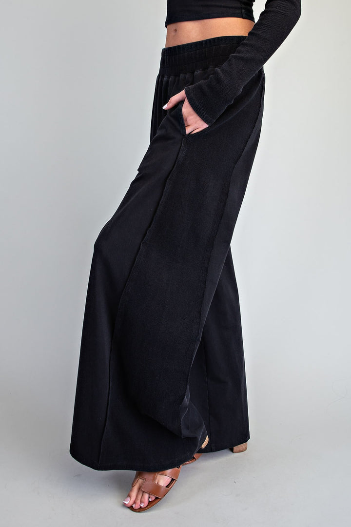 A person wearing black wide-leg draped pants with side pockets, paired with a black long-sleeve crop top and brown heeled sandals, standing against a light gray studio background.