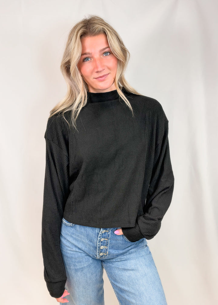 A woman with blonde hair wearing a black ribbed mock neck long-sleeve sweater and light blue jeans, standing against a white studio backdrop.