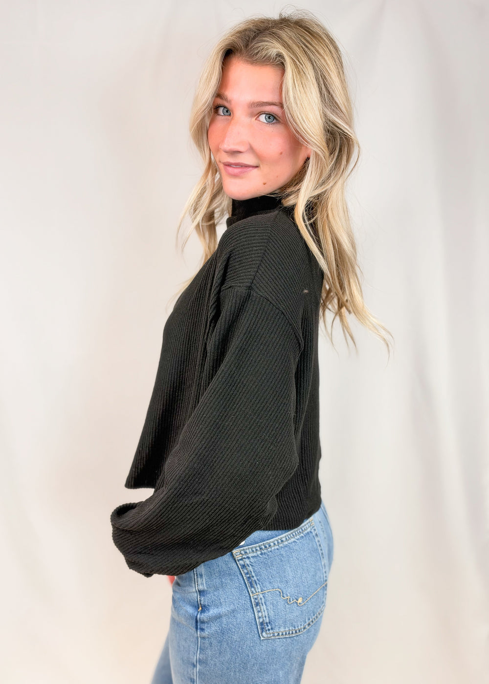 A woman with blonde hair wearing a black ribbed long-sleeve mock neck sweater and light blue jeans, posing against a white fabric backdrop.