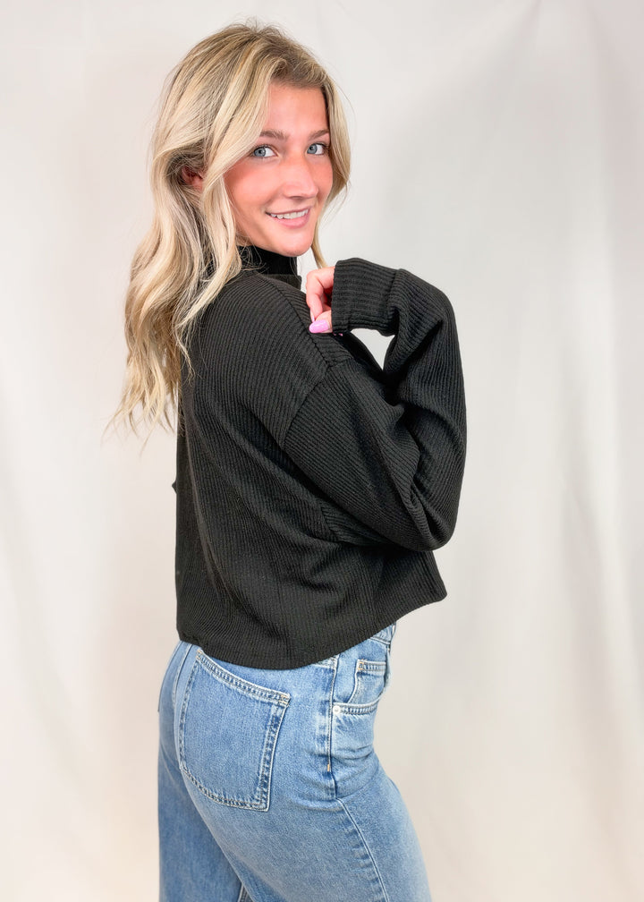 A woman with blonde hair wearing a black ribbed long-sleeve mock neck sweater and light blue jeans, posing against a white studio backdrop.