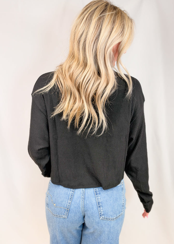 A woman viewed from behind wearing a black ribbed long-sleeve top and light blue jeans, standing against a plain white studio backdrop.
