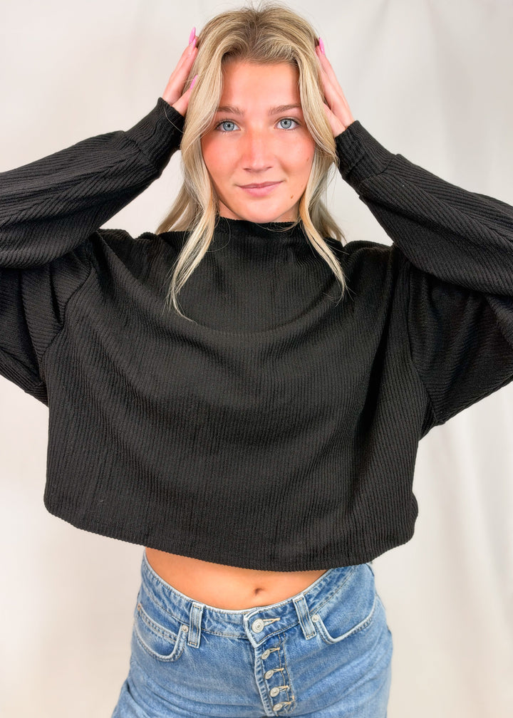 A woman wearing a black ribbed long-sleeve mock neck crop top and blue jeans, posing with her hands in her hair against a light neutral studio backdrop.