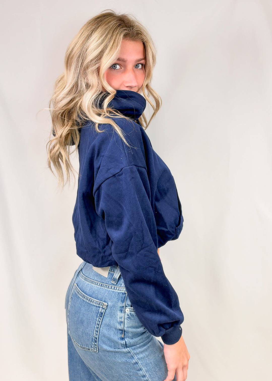 Woman shown from side view wearing cropped blue hooded sweatshirt with jeans on plain background