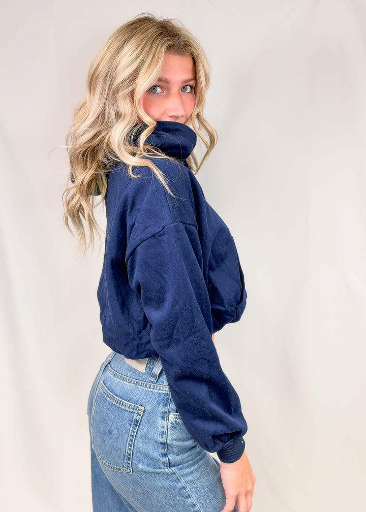 Woman shown from side view wearing cropped blue hooded sweatshirt with jeans on plain background