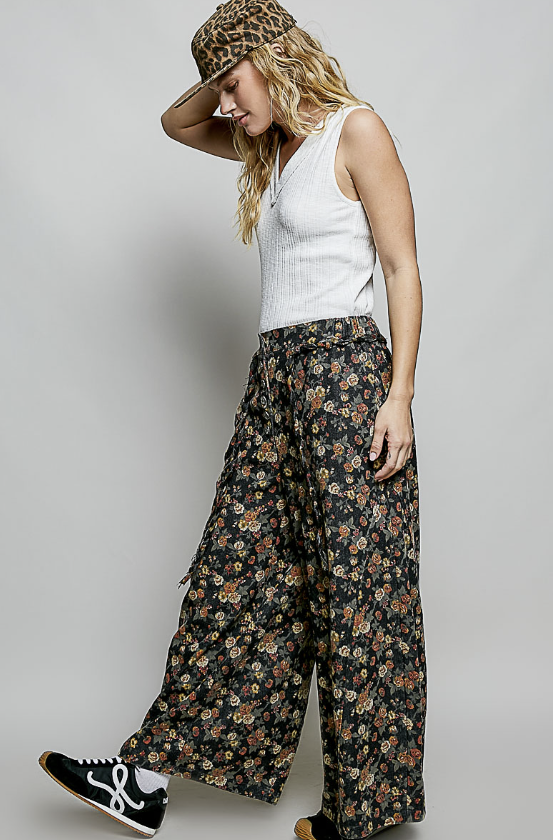 A woman posing in a studio wearing a leopard-print cap, a white ribbed tank top, wide-leg black floral pants, and black sneakers with white laces, set against a solid light gray background.