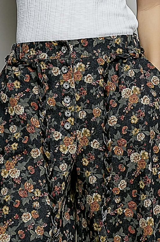 Close-up of a person wearing high-waisted, wide-leg trousers with a black base and a dense multicolor floral print in orange, yellow, cream, and green. A white ribbed top is partially visible at the waist.
