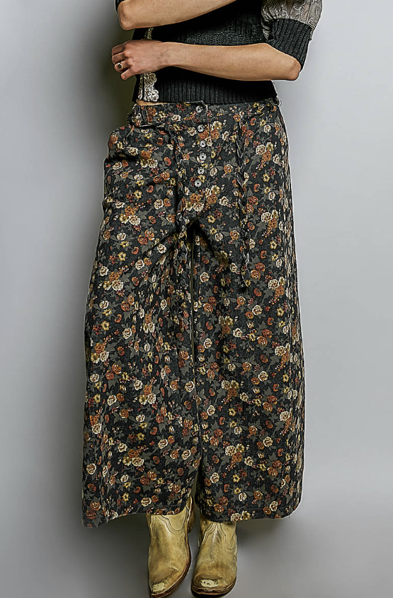 A person wearing wide-leg floral skirt pants in a dark gray/black base with a dense multicolor floral print, styled with a dark top and tan boots against a plain gray background.