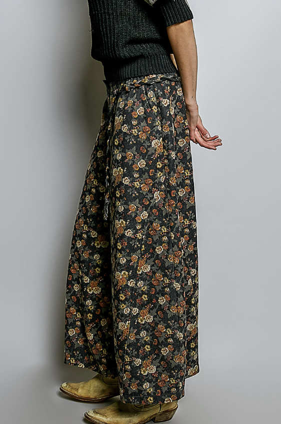 A person shown from the back wearing wide-leg floral pants with a dark charcoal base and small multicolor flowers, paired with a dark ribbed sweater and tan boots against a plain light gray studio background.